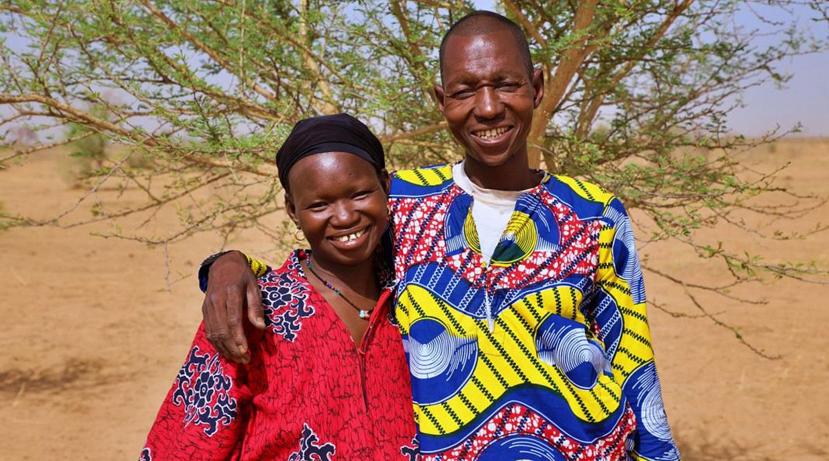 Afrika, Mali: Ein Ehepaar blickt lachend in die Kamera. Sie tragen beide bunte, traditionelle Kleider. Hinter ihnen steht ein grüner Baum.