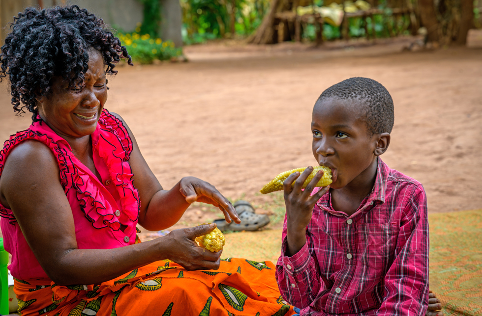 Eine Frau und ein Kind sitzen auf einer Matte und essen Maiskolben in Sambia.