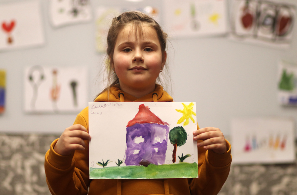 Ukraine: Ein Mädchen zeigt ein gemaltes Bild mit einem Haus, Garten und Sonne..