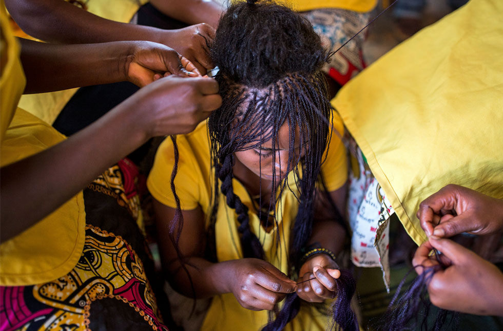 Demokratische Republik Kongo/DRK/DRC: Einige Frauen frisieren die Haare einer anderen Frau. Die Frauen tragen bunte, meist gelbe Kleider.