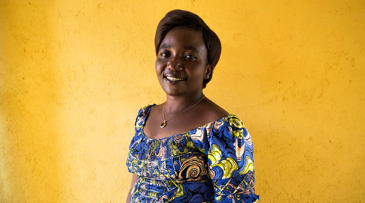 Demokratische Republik Kongo: Eine Frau in einem blaugemusterten Kleid steht lachend vor einer gelben Wand.