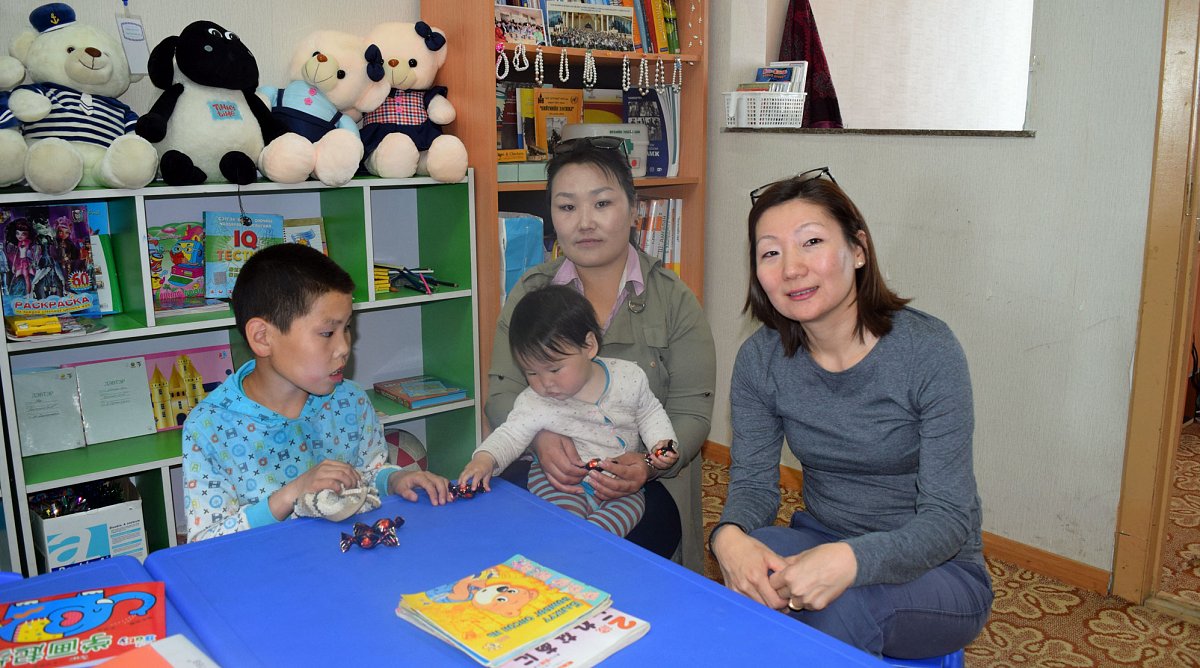 Mongolei: Zwei Frauen spielen mit zwei Kindern an einem kleinen blauen Tisch in einem Spielzimmer.
