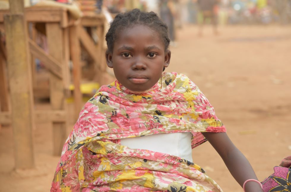 in junges Mädchen aus der Demorkatischen Republik Kongo mit einem geblümten Tuch um die Schultern sitzt auf einem Markt und schaut in die Kamera.