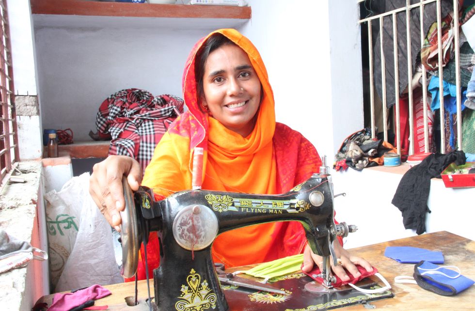 Ein Mädchen aus Bangladesch in einem orangenen Sari sitzt hinter einer Nähmaschine und näht  Mund-Nasenmasken.
