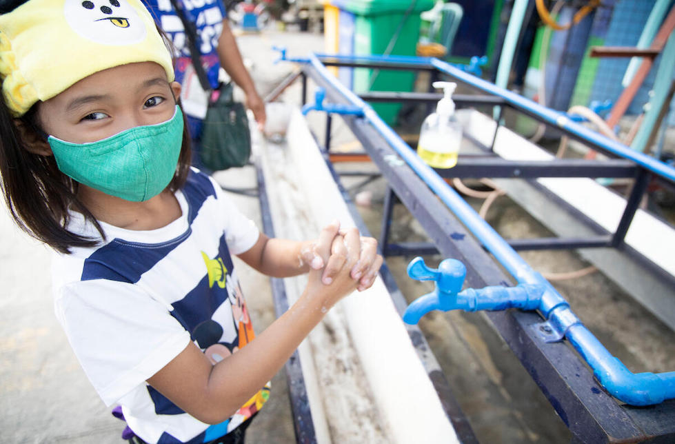 Philippinen: ein junges Mädchen mit Maske, steht an einer langen Handwaschanlage und seift sich die Hände unter einem Wasserhahn ein. Dabei blickt sie lächelnd in die Kamera. 