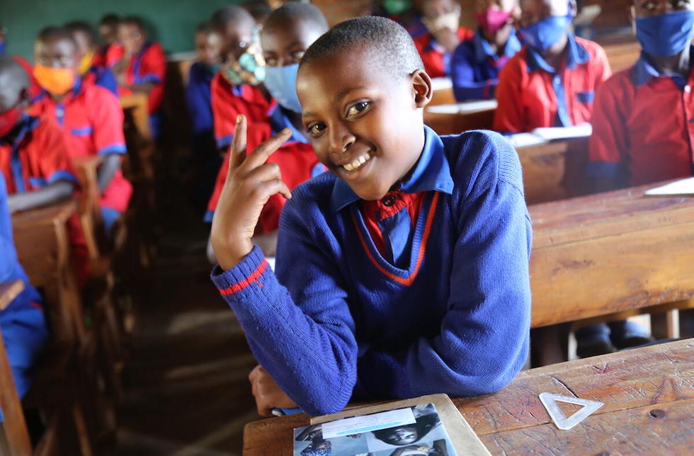 Ruanda: Ein junges Mädchen in Schuluniform sitzt im Klassenzimmer und macht ein Peace-Zeichen mit der Hand. In der Unschärfe hinter ihr sitzen ihre Mitschüler. 