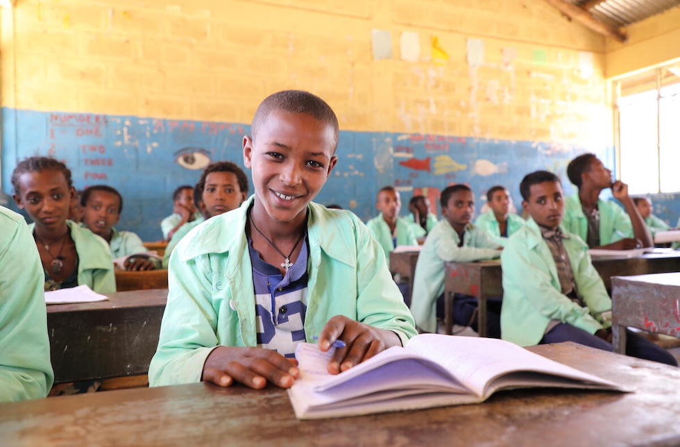 Äthiopien: Ein Junge sitzt in der Schulbank und blättert in einem Buch, das vor ihm liegt. Dabei lächelt er in die Kamera. Im Klassenzimmer hinter ihm sitzen seine Mitschüler.