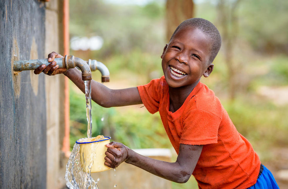 Kenia: Ein lachender Junge füllt an einem Wasserhahn seinen Becher mit Wasser auf.