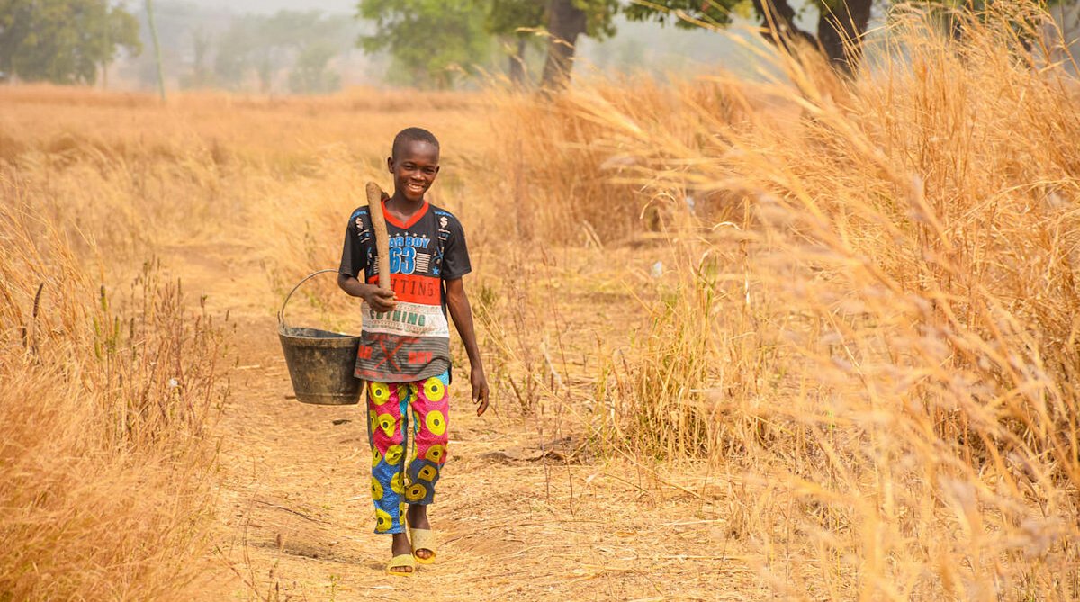 Ghana: Ein Junge in bunten Klamotten läuft mit einem Eimer im Arm einen trockenen Feldweg entlang.