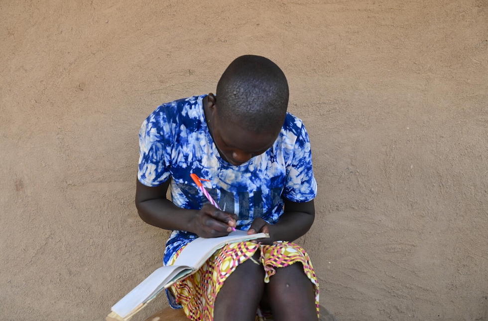 Kenia: Ein junges Mädchen sitzt an einer Wand und schreibt in ein Heft, dass auf ihren Beinen liegt.  