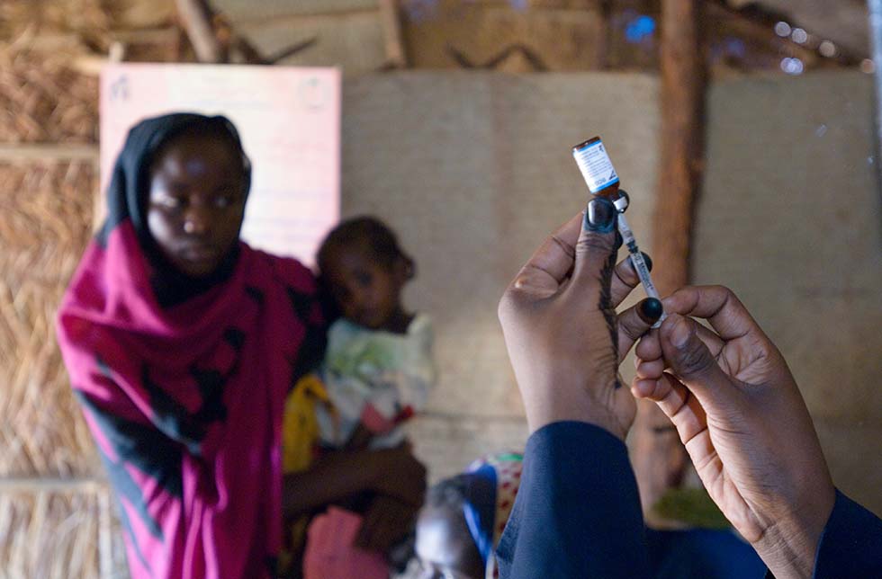 Sudan: Ein Paar Hände hält eine Spritze und Injektionsmittel, eine Mutter mit Kind schaut dabei zu.