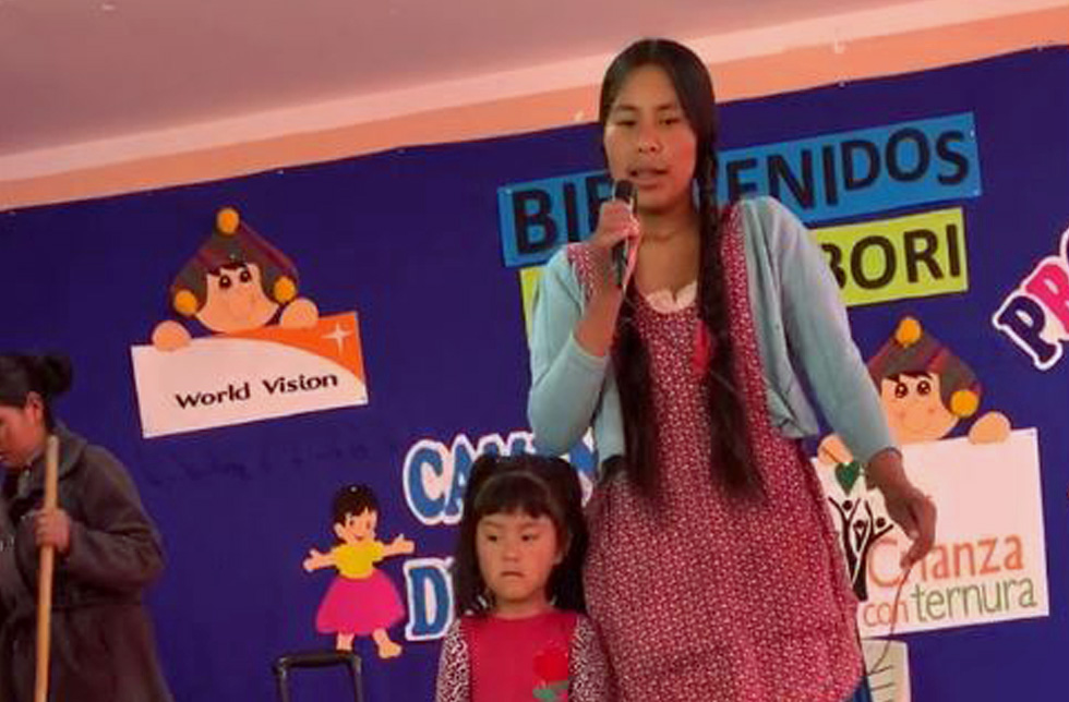 Eine junge Frau mit Zöpfen und Schürze steht auf einer Bühne am Mikrophon und spricht. Neben ihr ein kleines Mädchen. Im Hintergrund Plakate zum Thema Kinderschutz.