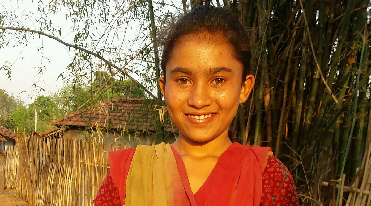 Indien: Ein Mädchen in einem roten Sari steht lächelnd vor einem Zaun aus Bambus.