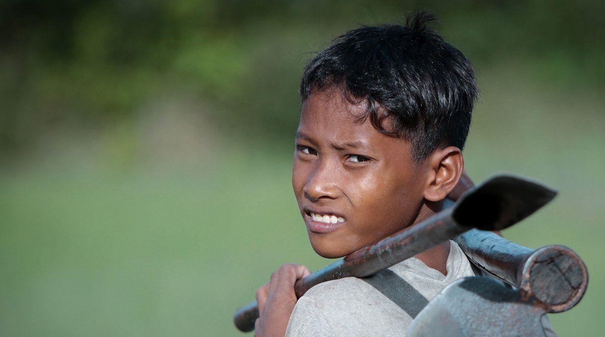Kambodscha: Ein Junge mit Werkzeug über den Schultern arbeiten.