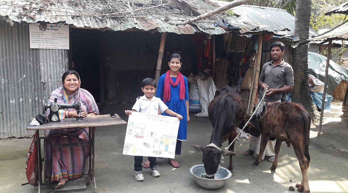 Bangladesch: Eine Frau sitzt an einer Nähmaschine vor ihrer Hütte. Neben ihr stehen ein junges Mädchen und ein Bub, der ein selbstgemaltes Bild hochhält. Rechts steht ein Mann der eine Kuh mit einem Kalb an einem Seil hält. Alle schauen fröhlich in die Ka