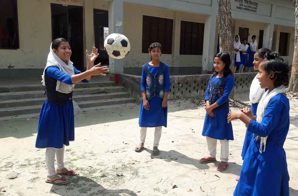 Bangladesch: Sechs Mädchen in blauen Schuluniformen stehen im Kreis vor einem Klassenzimmer und werfen sich einen Ball zu.