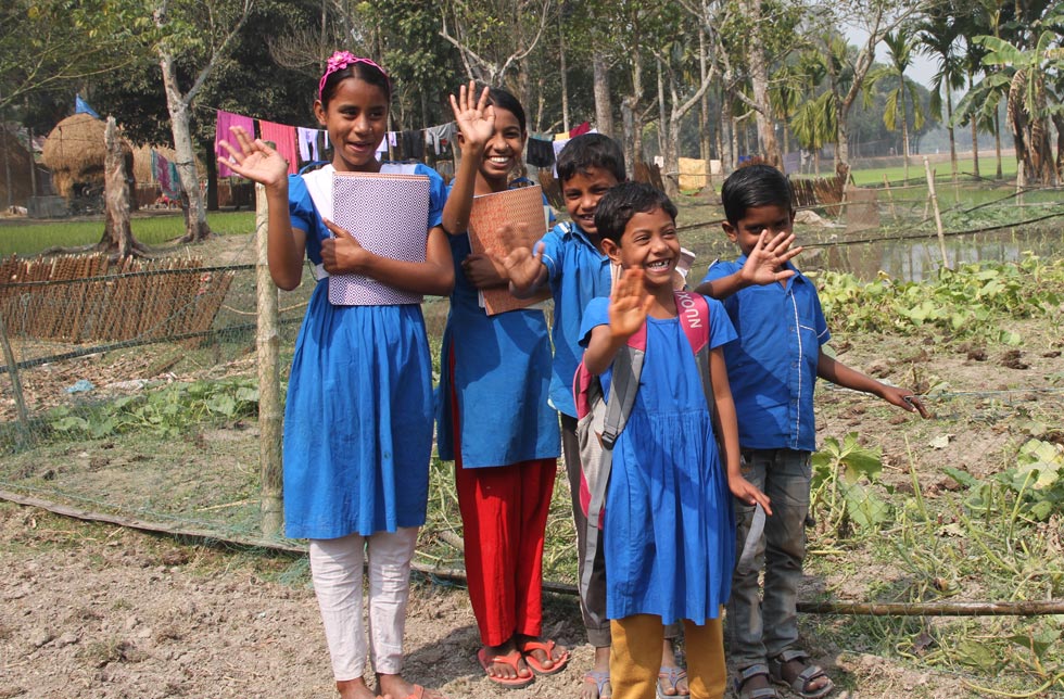 Fünf Kinder aus Bangladesch mit einfachen, königsblauen Schuluniformen winken jemandem zu.