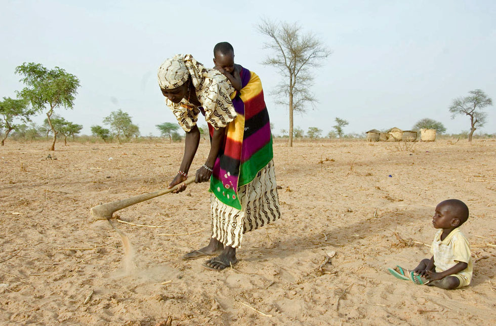 Niger: Eine Frau in bunter Kleidung steht auf einem vertrockneten Feld und gräbt mit einer einfachen Holzhacke den Boden um. Auf dem Rücken trägt sie einen Säugling, auf dem Boden sitzt ein Kleinkind. 