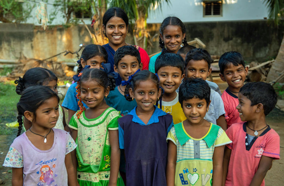 Indien: Eine Gruppe Mädchen lacht in die Kamera. Im Hintergrund eine junge Frau.