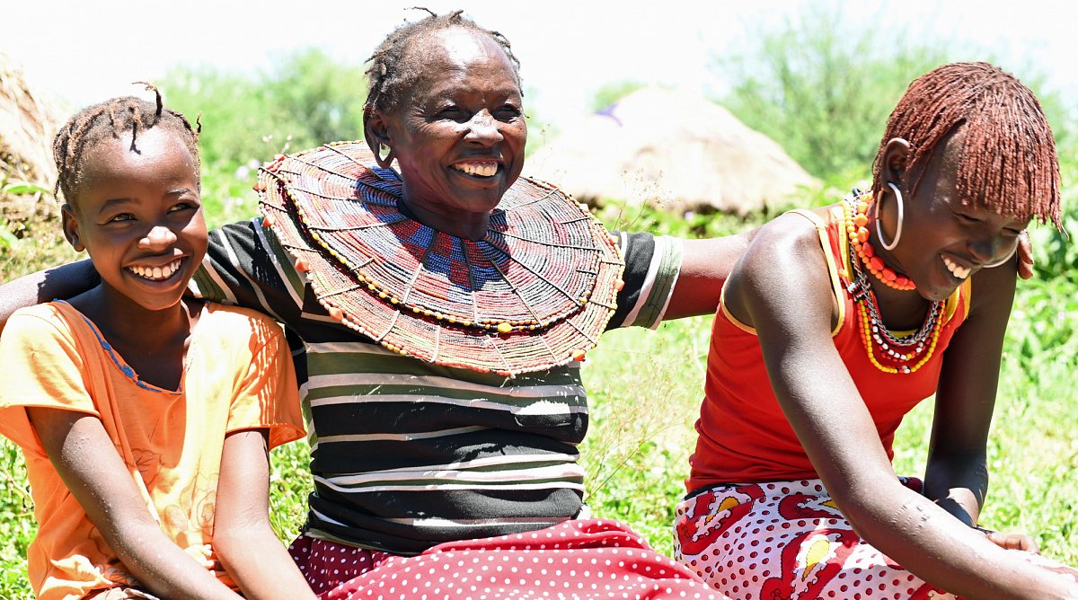 Kenia: Eine Frau legt lachend ihre arme um zwei Mädchen, die ebenfalls lachen.