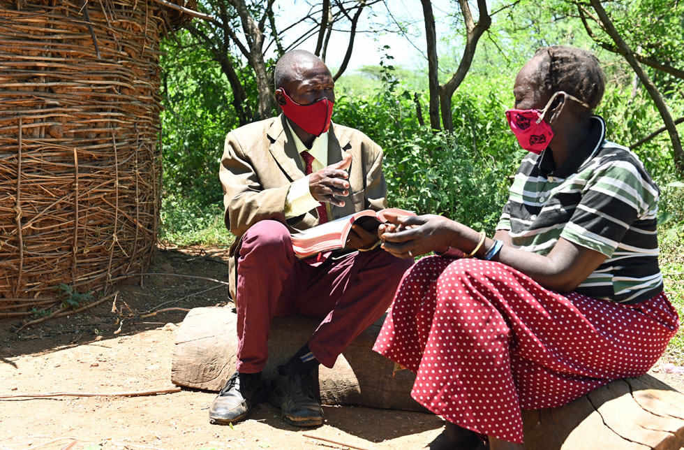 Kenia: Ein Mann und eine Frau sitzen auf Holzstrünken vor einer traditionellen Hütte und reden miteinander.