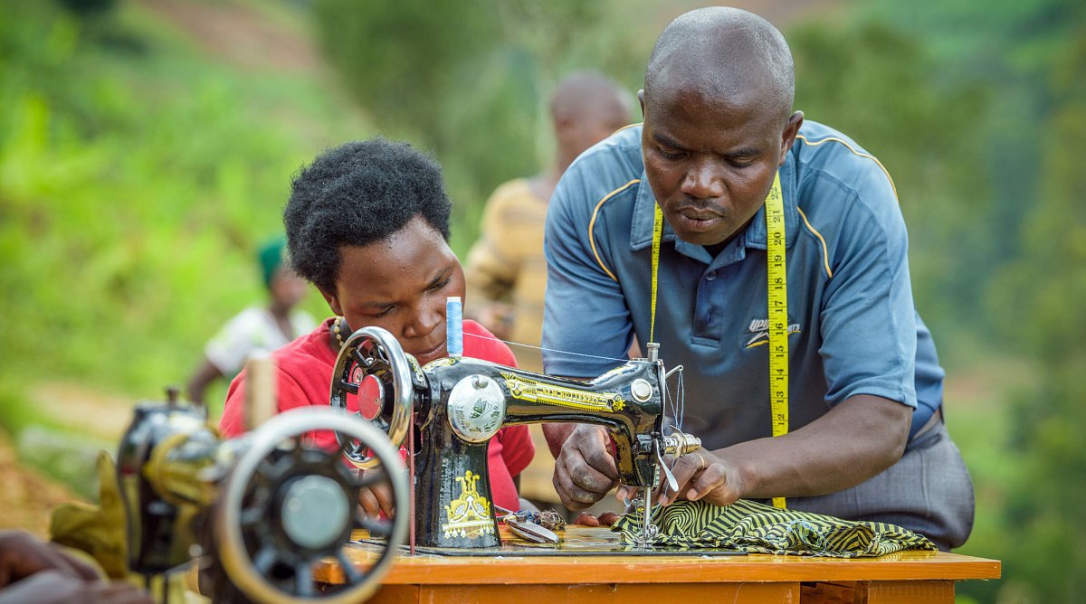 Ruanda: Ein Mann erklärt einer Frau an einer Nähmaschine, wie man richtig näht.