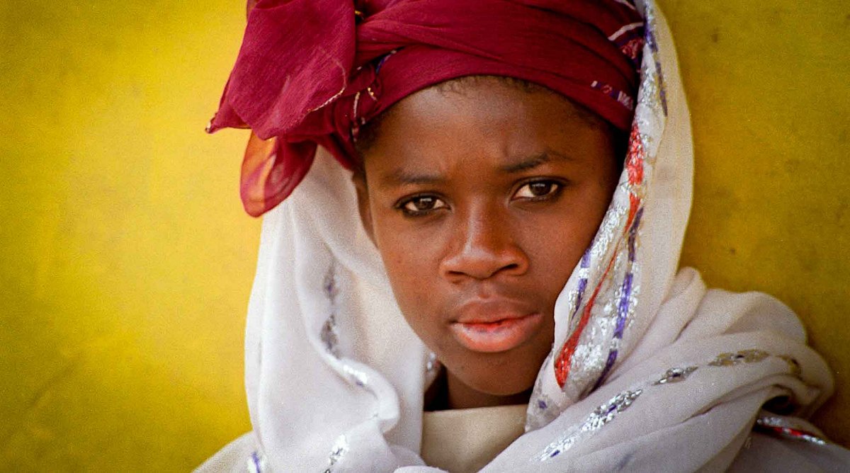 Ein junges Mädchen aus Ghana mit rotem Kopftuch und weissem Schal lehnt an eine gelbe Wand.