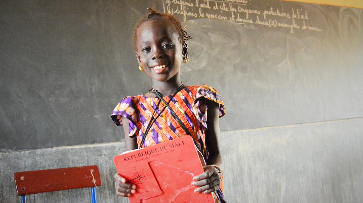 Ein kleines Mädchen steht in Mali in der Schule vor der Tafel und lächelt in die Kamera.