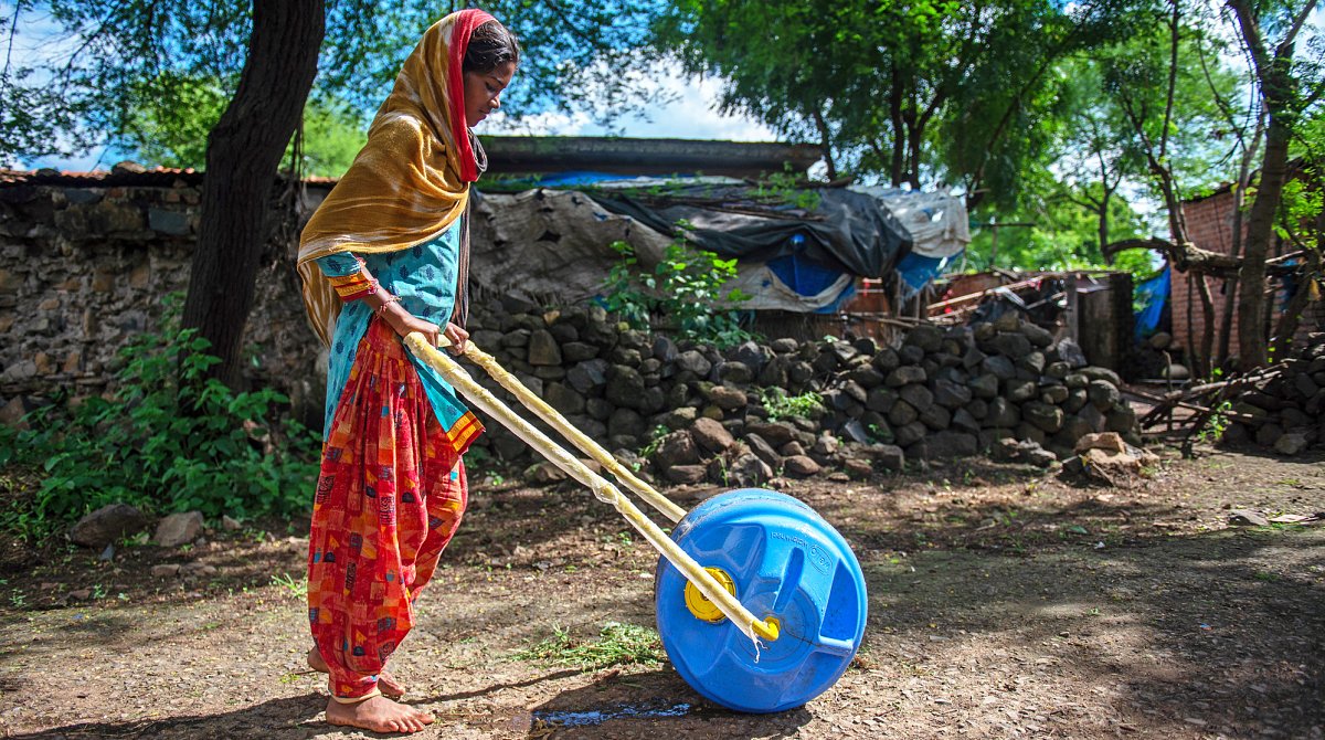 Indien: Mädchen stösst ein mit Wasser gefülltes Wasserrad aus blauem Kunststoff vor sich her.