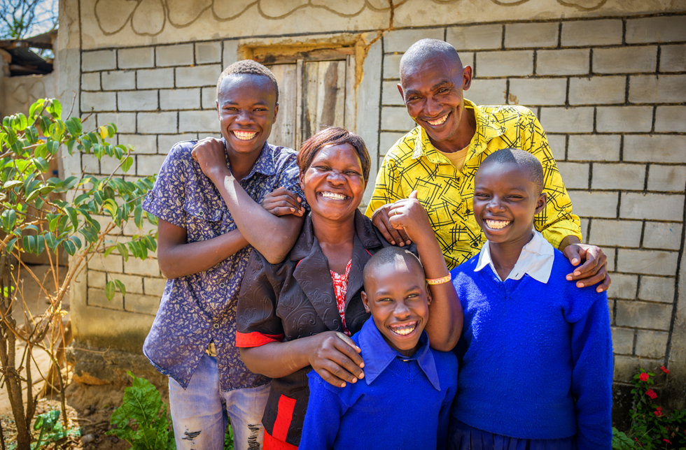 eine fünf-köpfige Familie steht fröhlich lachend vor ihrem Haus in Kenia