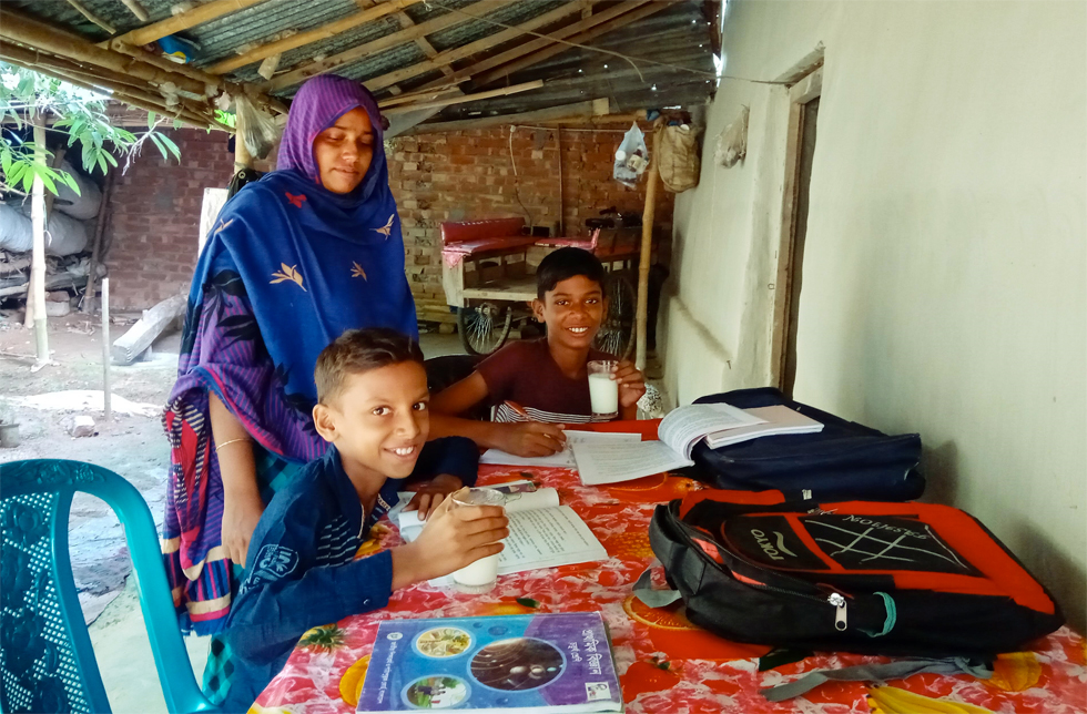 Bangladesch: Zwei Jungen sitzen an einem Tisch und lernen, ihre Mutter steht dabei.