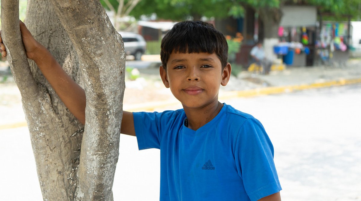 Honduras: Junge lehnt an einen Baumstamm und blickt direkt in die Kamera.