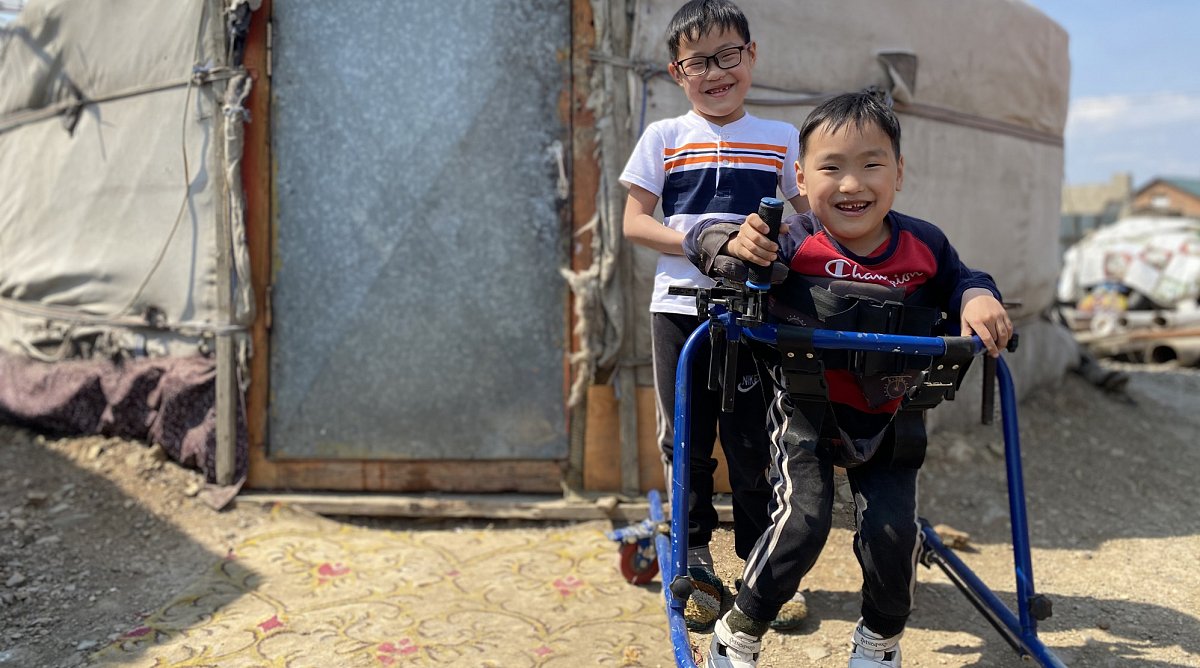 Mongolei: Ein Junge mit einer Behinderung steht in seinem Gehtrainer, sein Bruder steht hinter ihm.