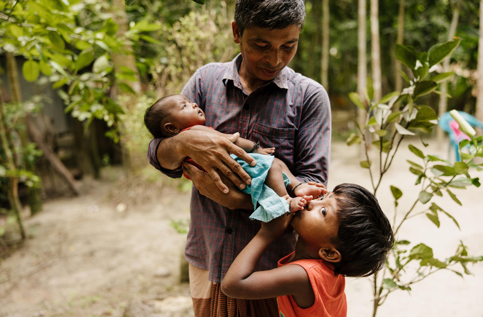 Bangladesch: Ein Vater hält einen Säugling im Arm, der von seinem älteren Geschwister die kleinen Füsse geküsst bekommt.