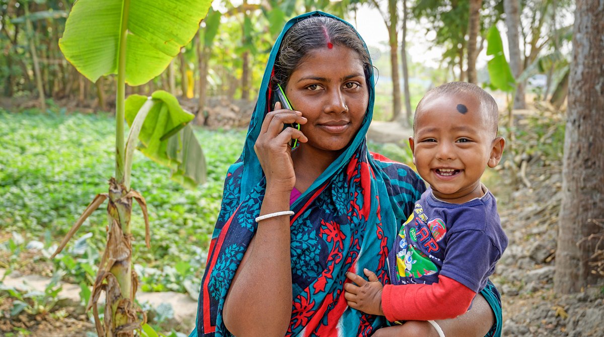 Eine Mutter in Bangladesch hält ihr fröhlich lächelndes Kleinkind auf dem Arm und telefoniert mit dem Mobiltelefon.