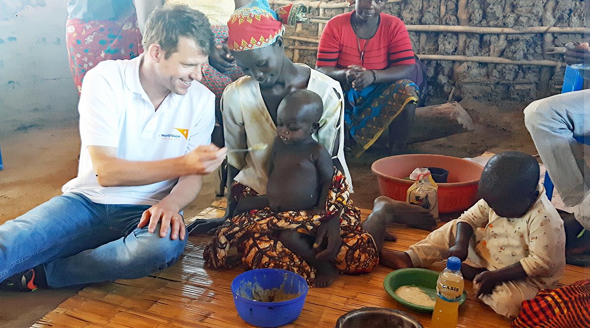 Der Länderverantwortliche Mosambik des Kinderhilfswerks World Vision Schweiz sitzt in einer Lehmhütte am Boden und füttert ein Kleinkind.