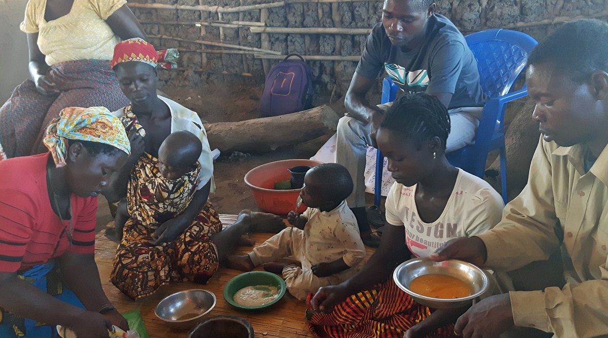 Frauen, Männer und Kinder sitzen teilweise auf einer Bodenmatte und bereiten gemeinsam ein gesundes Essen mit lokalen Zutaten zu.