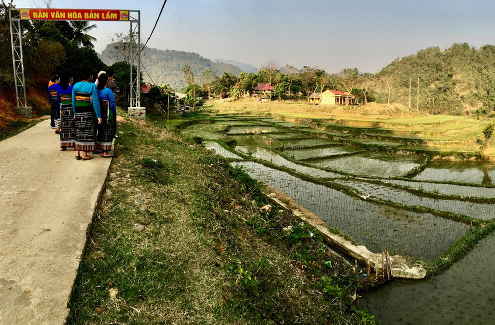 Vietnam: Reisfelder mit mehreren Terrassen, am Strassenrand Frauen, im Hintergrund ein Dorf.