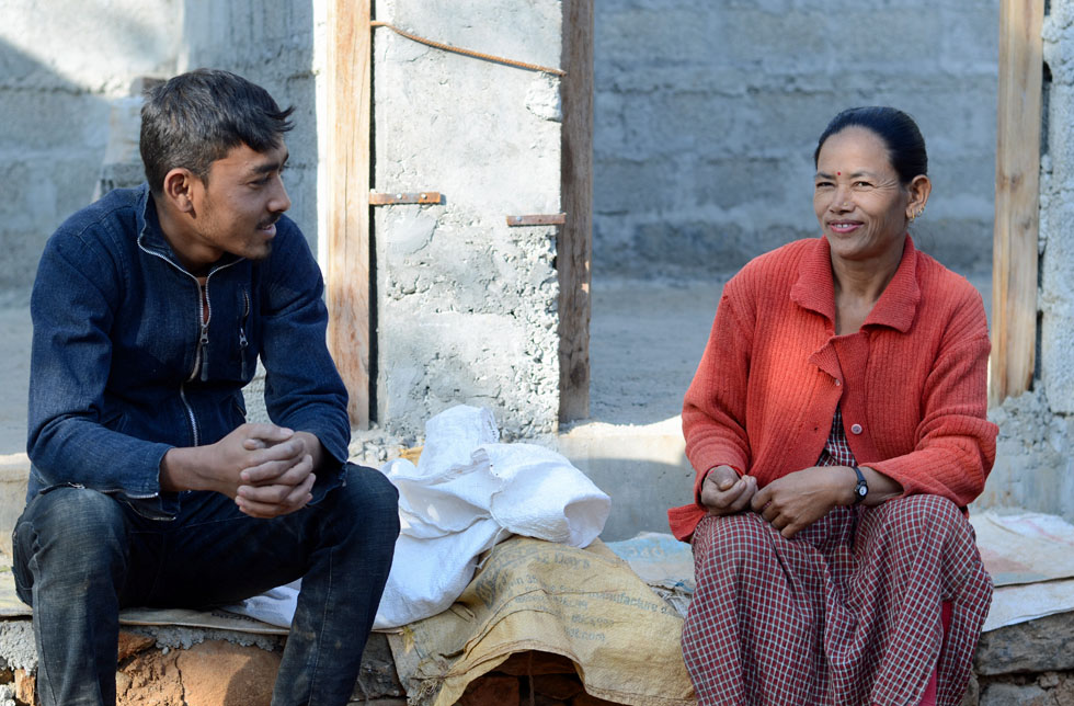 Nepal: Eine Frau in einer roten Strickjacke und kariertem Kleid und ein junger Mann in Jeans und blauer Jacke sitzen gemeinsam vor einem unfertigen Haus und schauen sich an.