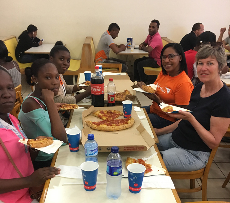 Dominikanische Republik: Eine Familie (Mutter, Tochter, Sohn) teilen sich mit der Schweizer Patin und einer lokalen World Vision-Mitarbeiterin eine grosse Pizza. Alle sitzen um einen Tisch herum.