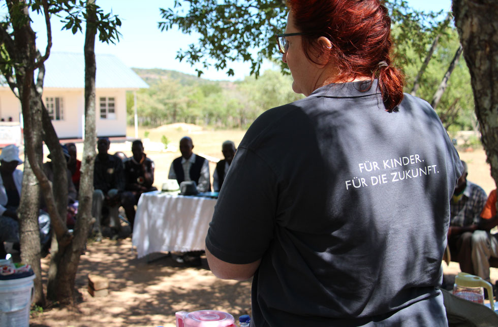 Simbabwe: Eine Frau mit roten Haaren steht vor einer versammelten Runde mit dem Rücken zur Kamera. Auf ihrem World Vision-T-Shirt steht: Für Kinder. Für die Zukunft.