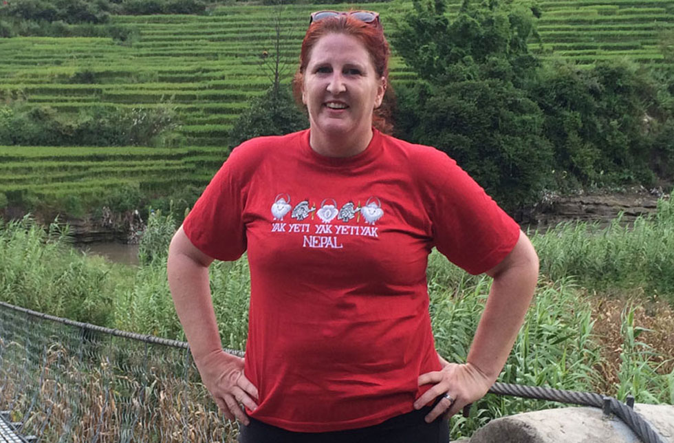 Nepal: Eine Frau mit roten Haaren und einem roten T-Shirt steht vor einer Hängebrücke. Im Hintergrund sind Reisterassen zu erkennen.