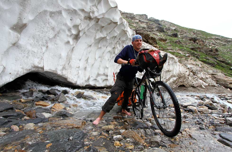 Himalaya: Ein erschöpfter Biker mit seinem bepackten Mountainbike barfuss  vor einem Eisblock
