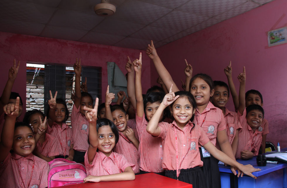 Bangladesch: Die Mädchen und Jungs einer Schulklasse strecken eifrig auf. Sie tragen alle rosa Schuluniformen und blicken lachend in die Kamera.