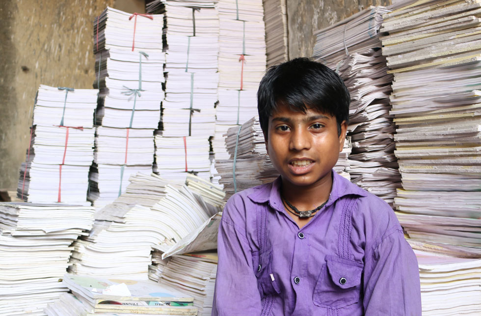 Indien: Ein Junge in einem violetten Hemd sitzt vor einigen Stapeln Papier.