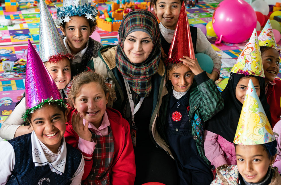 Mossul, Irak: Eine Gruppe Kinder und eine Frau sitzen mit bunten Hüten und lachend in der Kinderschutzzone von World Vision.