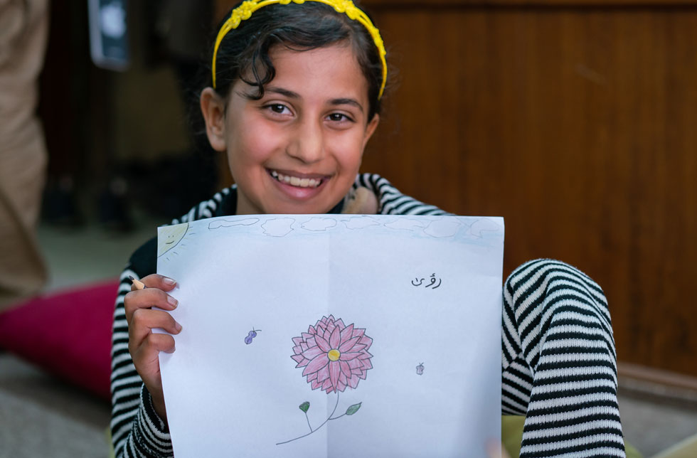Mossul, Irak: Ein Mädchen blickt lachend in die Kamera. In ihrer Hand hält sie eine selbstgemalte Zeichnung von einer rosaroten Blume.