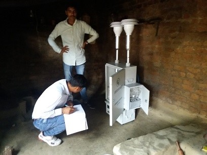 Indien: Im inneren einer verrauchten Hütte messen zwei Männer in Jeans und weissen Hemden mit einem Emissionsmessgerät die Luftqualität.