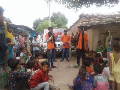 Indien: Drei junge Männer in orangen Shirts mit Mikrophonen tragen auf der Strasse zwischen 2 strohgedeckten Hütten etwas vor. Viele Frauen, Männer und Kinder sitzen davor auf dem Boden uns hören zu.