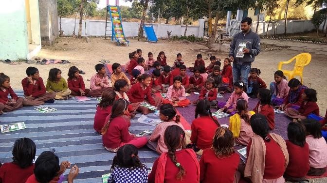 Indien: Eine Gruppe Kinder in Schuluniformen sitzt im Kreis auf einer ausgebreiteten Plane und sieht einem Lehrer zu, der etwas erklärt.
